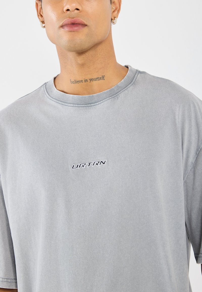 Szara bawełniana koszulka z luźnym krojem, z okrągłym dekoltem i haftowanym logo "URTRN" na piersi. Miękka tekstura, minimalistyczny design.