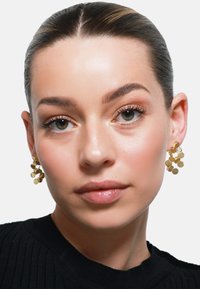 Guldtonede øreringe med teksturerede cirkulære skiver arrangeret i et klynge-design, båret af en model med en neutral makeuppalette.
