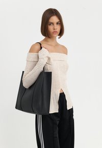 Černá kožená taška s hladkým povrchem a vyvýšenými švy, kterou drží osoba v lehkém ribovaném topu a černých teplákových kalhotách s bílými pruhy.