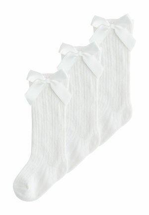 Trys poros baltų raštuotų kelio aukščio kojinių su didelėmis baltomis satino kaspinais viršuje, išdėstytos vienos ant kitų ant balto fono.