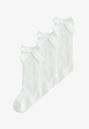 Trois paires de chaussettes blanches à côtes montant jusqu'aux genoux, chacune ornée de grands nœuds en satin blanc en haut, disposées en chevauchement sur un fond blanc.