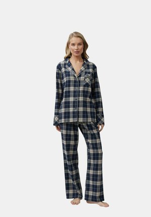 Donna in piedi a piedi nudi che indossa una camicia da pigiama a maniche lunghe con bottoni, a quadri blu navy e bianchi, e pantaloni da pigiama abbinati, larghi.