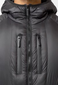 Čierna puffa bunda s predným zipsom, dvoma zipsovými vreckami na hrudi a kapucňou. Materiál vyzerá hladko s lesklým povrchom.