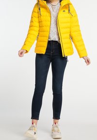 Gul puffjacka med en figursydd design, dragkedjefickor och huva. Bärs över en ljus mönstrad topp och matchas med mörka tighta jeans och flerfärgade sneakers.