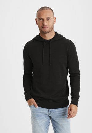 Schwarzer Baumwollhoodie mit langen Ärmeln, einer Kapuze mit Kordelzug und einem engen Saum, getragen über hellblauen Jeans. Schlichtes und funktionales Design.