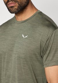 Oliivroheline sporditee, mis on valmistatud kergest tekstuuriga kangast. Omab lühikeseid varrukaid ja rinnal on väike valge logo.