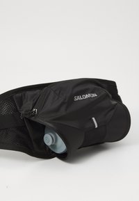 Ce sac ceinture noir est fabriqué en tissu durable et possède une poche pour bouteille d'eau. Il dispose d'une fermeture éclair et d'accents en mesh pour une meilleure respirabilité.