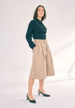 Camicia nera con bottoni, con maniche lunghe, abbinata a culotte beige a vita alta e décolleté neri con cinturino. Design semplice e strutturato.