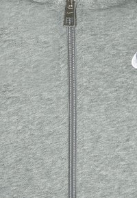 Grå zip-up sweatshirt med texturerad tyg, med en metallisk dragkedja och en vit logotypdetalj på vänster sida.