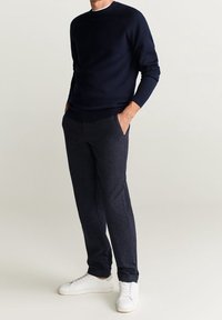 Pull en maille bleu marine avec col et poignets côtelés, associé à un pantalon foncé à motifs. Des baskets blanches complètent la tenue.