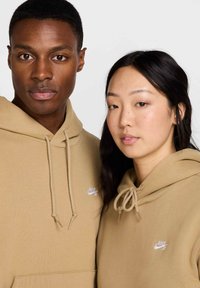 Deux individus portent des sweat-shirts à capuche beige assortis en tissu doux. Chaque hoodie arbore un logo Nike blanc sur la poitrine gauche et des cordons de serrage.