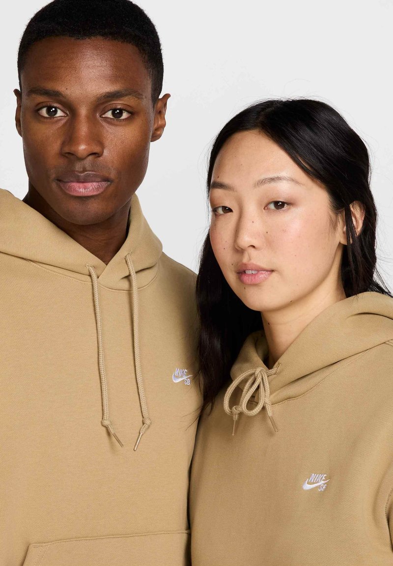 Dwie osoby noszą dopasowane beżowe bluzy z kapturem wykonane z miękkiego materiału. Każda bluza posiada białe logo Nike na lewej piersi oraz sznurki.