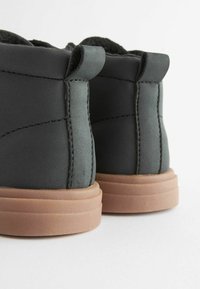 Stivaletti neri sintetici con una superficie liscia, suole in gomma marrone chiaro a contrasto e dettagli cuciti discreti sui lati.
