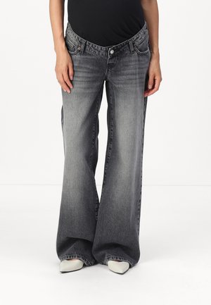 Person iført højttaljede, vidde, falmede sorte jeans med hvide spidse sko, stående mod en ensartet hvid baggrund.