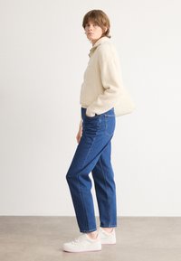 Kremfarget ribbet genser med krage, kombinert med høyhårede blå denimjeans. Modellen har på seg hvite joggesko med rosa detaljer.