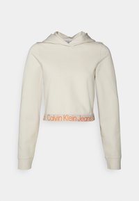 Skrojona beżowa bluza z kapturem wykonana z miękkiego materiału, z długimi rękawami, kieszenią z przodu oraz pomarańczowym logo "Calvin Klein Jeans" na dolnym brzegu.