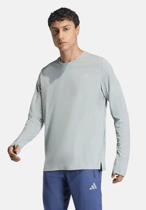 Homme portant une chemise à manches longues gris clair et un pantalon de sport bleu avec le logo Adidas, debout devant un fond blanc uni.