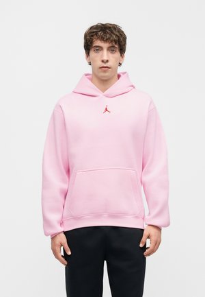 Młody mężczyzna w jasnoróżowej bluzie z kapturem z małym czerwonym logo Jumpman i czarnych spodniach, stojący na jednolitym jasnym tle.