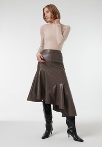 Jupe évasée en cuir brun avec un ourlet asymétrique, associée à un haut beige à manches longues et des bottes noires à talons aiguilles arrivant au genou.