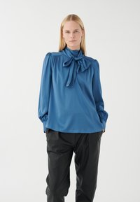 Blaue Bluse mit langen Ärmeln und einer Schleife am Hals, plissierten Schultern und glattem Stoff. Kombiniert mit lockeren schwarzen Hosen.