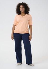 Lichtperzikkleurige korte mouwen V-hals top, gedragen met donkerblauwe flared jeans. De outfit heeft een ontspannen pasvorm en een gladde stofstructuur.