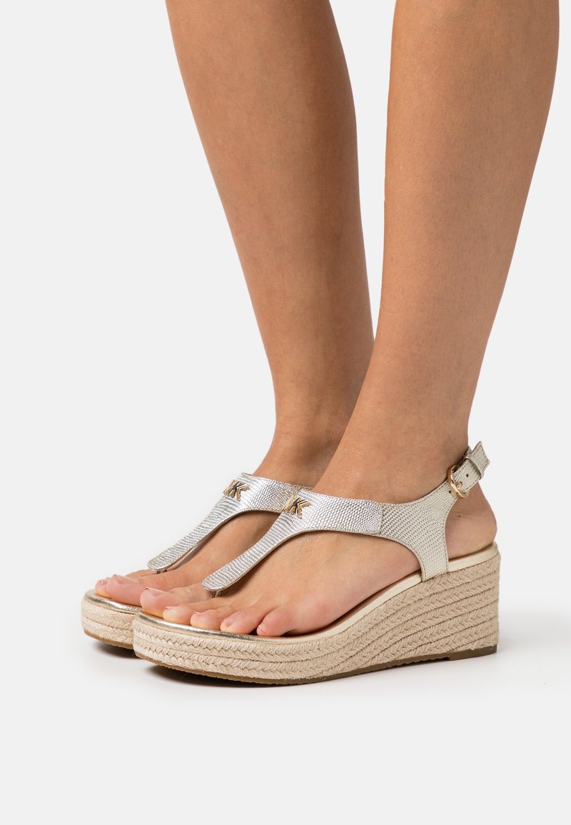 Pieds chaussés de sandales compensées argentées métalliques avec brides à la cheville et semelles en corde tressée sur fond blanc.