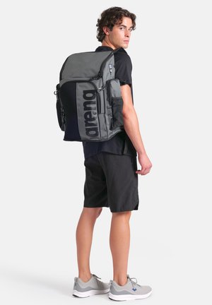 Gray backpack with a large black logo, multiple compartments, mesh side pockets, and adjustable padded straps, worn by a person in black shorts.