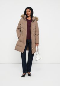 Mujer vestida con un abrigo tipo puffer con capucha beige y ribete de piel, top burdeos, jeans oscuros, tacones negros, sosteniendo un pequeño bolso blanco contra un fondo sencillo.