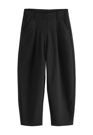 Ruka v vrecku na čiernych nohaviciach s vysokým pásom, nosených so zastrčenou bielou košeľou na neutrálne pozadí.