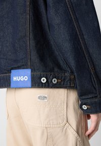 Tamni traper jakna s plavom oznakom "HUGO", srebrnim kopčama i kontrastnim narančastim šavovima. Bež hlače s oznakom "JACK & JONES".