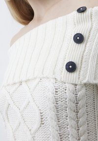 Schulterfreier cremefarbener Strickpullover mit strukturiertem Rippstrick, Zopfmuster und drei dunklen Knöpfen vorne.