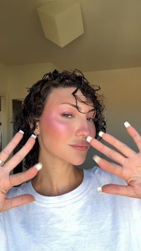 Cheveux bouclés, blush rose clair sur les joues, couleur de lèvres neutre, t-shirt blanc à col rond, mains avec des ongles longs et vernis blancs levées devant.