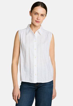 Femme aux cheveux foncés portant une chemise blanche sans manches boutonnée et un jean bleu sur un fond clair uni.