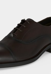 Chaussure habillée Oxford en cuir marron foncé avec lacets fermés, bout droit et petit talon sur un fond clair uni.