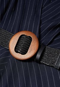 Musta kudottu vyö pyöreällä puukannella, jossa on leikkauskuvio. Taustalla on laivastonsininen raidallinen kangas.