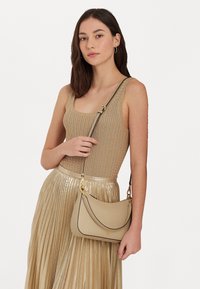 Beige axelväska med gyllene detaljer, rundad form, mjuk textur och ett avtagbart handtag, hålls av en person som bär en ribbad topp och en veckad kjol.