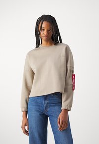 Beige cropped sweatshirt with ribbed neckline, long sleeves, and elastic cuffs. Features a red tag on the sleeve and soft fabric texture.