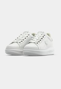 Paire de baskets basses en cuir blanc avec lacets, logo embossé sur le côté et semelles épaisses blanches sur un fond uni.