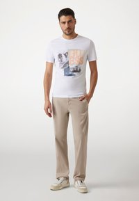 Biela bavlnená T-shirt s grafickým dizajnom, na prednej strane s nápisom "GUESS" v oranžovej a modrej farbe, v kombinácii s béžovými nohavicami a bielymi teniskami.