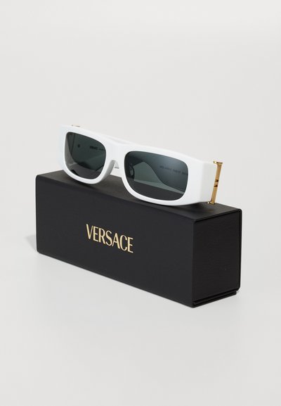 Lunettes de soleil rectangulaires blanches avec des verres foncés, accentuées de doré sur les charnières, présentées sur une boîte noire marquée "VERSACE" en lettres dorées.