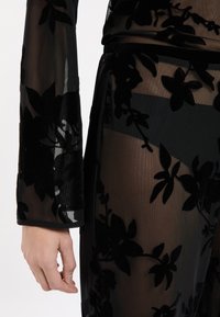 Hand met licht gebogen vingers naast doorschijnende zwarte bloemenkant stof die arm en been bedekt, met een glimp van de huid eronder en details van de tailleband.