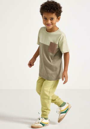 Grøn og brun farveopdelt t-shirt med en brystlomme, kombineret med teksturerede lysegrønne joggers og beige sneakers med grønne og gule accenter.