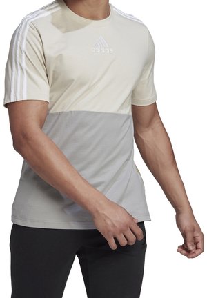 Man draagt een tweekleurig beige en grijs Adidas T-shirt met korte mouwen, witte strepen op de schouders en zwarte broek, tegen een witte achtergrond.