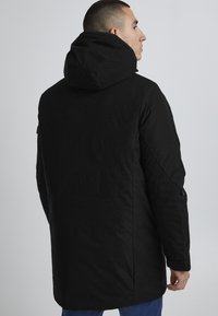 Cămașă neagră căptușită cu o glugă mare, buzunare laterale cu fermoar și croială dreaptă. Prezintă o textură netedă și un design minimalist.