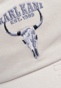 Fehér pamut sapka, amelyen egy hímezett bika koponya látható szürke és fekete részletekkel, a tervezés fölött pedig a "KARL KANI EST. 1989" felirat.