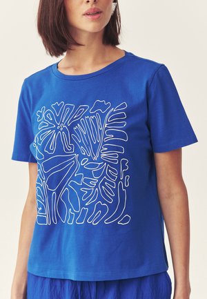 Kvinde iført en blå kortærmet skjorte med hvid abstrakt botanisk linjekunst på forsiden, kombineret med matchende blå bukser.