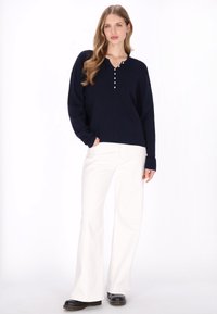 Pulover bleumarin tricotat cu decolteu în V și detalii cu nasturi, asortat cu pantaloni evazați albi și pantofi negri. Textură moale și croială lejeră.