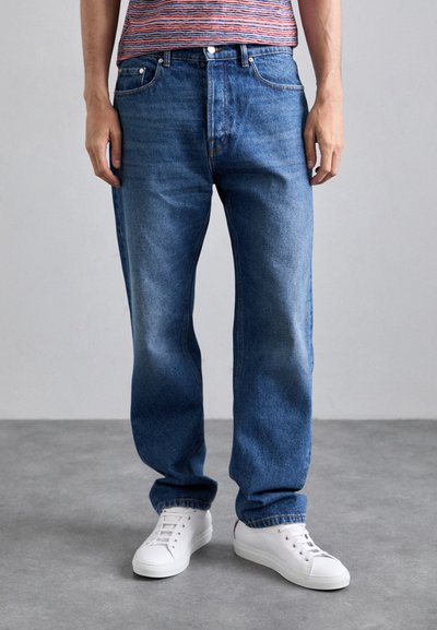 Jean en denim bleu avec une coupe droite, texture délavée, design à cinq poches et finitions en métal argenté, associés à des baskets blanches.