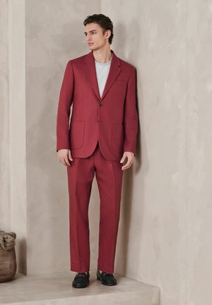 Jeune homme portant un costume bordeaux sur mesure avec une chemise rayée blanche et grise, debout contre un mur texturé beige sur une plateforme surélevée.