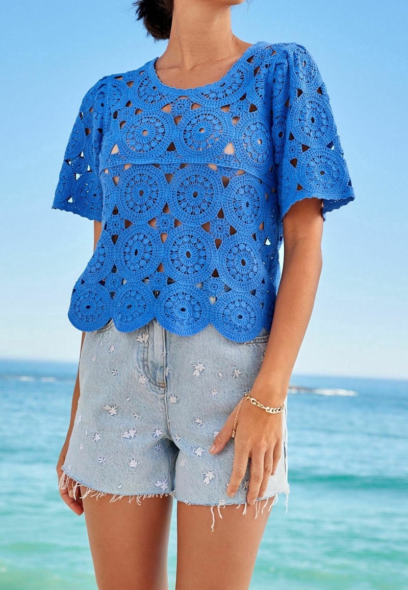 Femme portant un haut bleu crocheté à manches courtes et un short en denim clair avec de petits motifs floraux blancs au bord de l'océan.